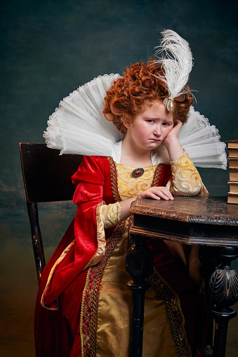 Portrait of little red-headed girl in costume of royal person sitting in tears isolated over dark green background. Concept of historical remake, comparison of eras, medieval fashion, emotions, queen