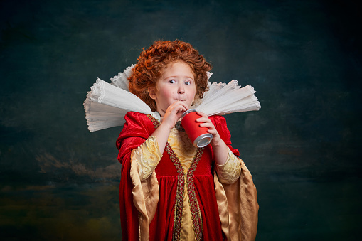 Portrait of little red-headed girl in costume of royal person drinking soda isolated over dark green background. Concept of historical remake, comparison of eras, medieval fashion, emotions, queen