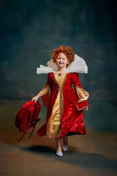 Photo of Portrait of little red-headed girl, child in costume of royal person isolated over dark green background. School time