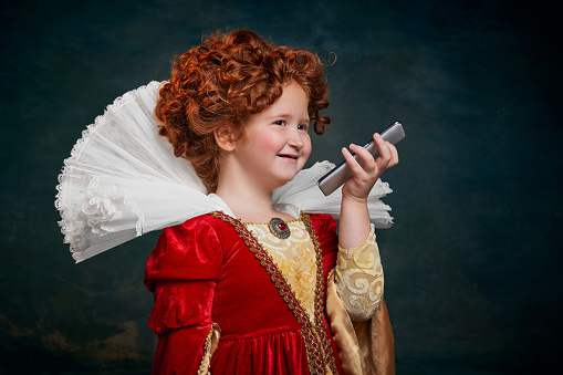 Portrait of little red-headed girl, royal person recording voice message on phone isolated on dark green background. Concept of historical remake, comparison of eras, medieval fashion, emotions, queen