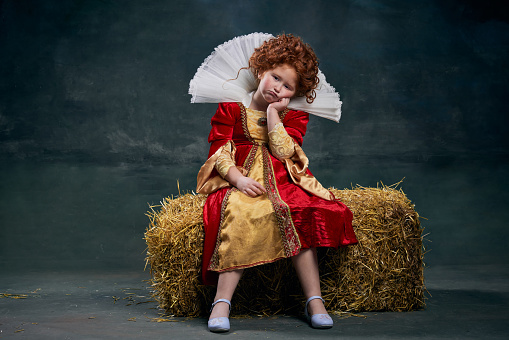 Portrait of red-headed child in image of royal person sitting on hay with boring face over dark green background. Concept of historical remake, comparison of eras, medieval fashion, emotions, queen