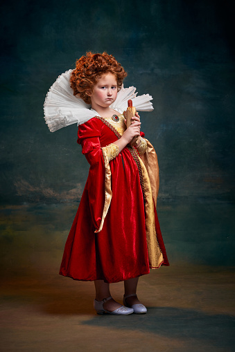 Little red-headed girl in image of royal person with serios face and hot-dog isolated over dark green background. Concept of historical remake, comparison of eras, medieval fashion, emotions, queen
