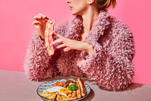 Stylish woman in pink furry coat eating american breakfast with fried eggs, sausages and bacon over pink background. Vintage, retro style. Food pop art. Complementary colors. Copy space for ad, text