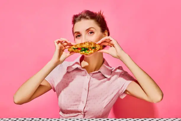 Photo of Woman eating freshly baked croissant with vegetables over pink background.
