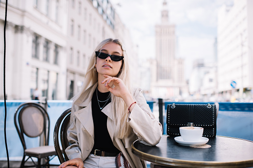 Young well dressed female with blond hair in sunglasses sitting in outdoor cafe in sunny day and waiting for meeting