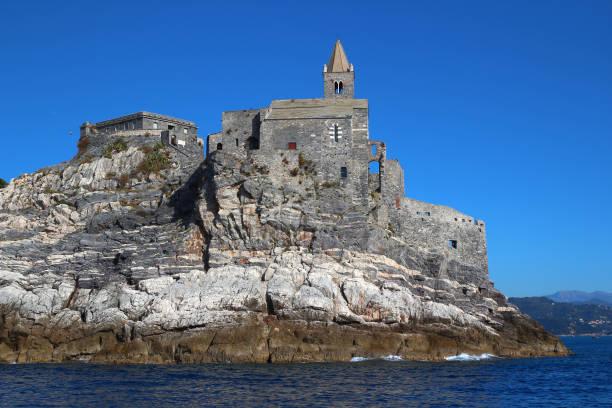 From the sea, view of the Church of San Pietro de Porto Venere on its rocky outcrop (Cinque Terre, Liguria, Italy) San Pietro is the first historical monument seen by the visitor upon arriving to Porto Venere by boat. It looks like a castle but it is only a church dating from the 13th century. Its façade has the traditional bands of the Ligurian style of the time. The church occupies an exceptional site, perched on a cape that juts out over the Mediterranean. church of san pietro photos stock pictures, royalty-free photos & images