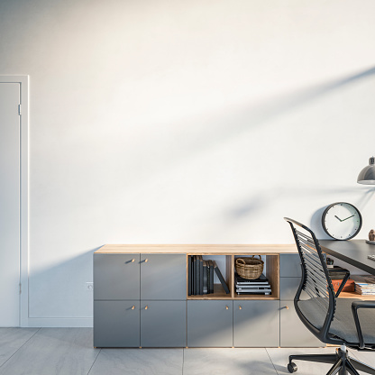 Home office - front view of a  cabinet and work desk with computer, equipment, and decoration (a clock, a printer,  books, a basket, lamp, chair, magazines, a door) on the gray tiled floor in front of an empty 
 white plaster wall with copy space.  A peek at a large and wide window over the work desk illuminates the desk. 3D rendered image.