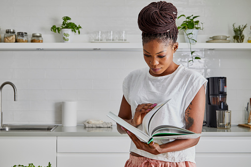 A beautiful black young adult woman in the kitchen, reading recipe book, for healthy breakfast prepared at the kitchen table, representing healthy lifestyle, an image with a large copy space