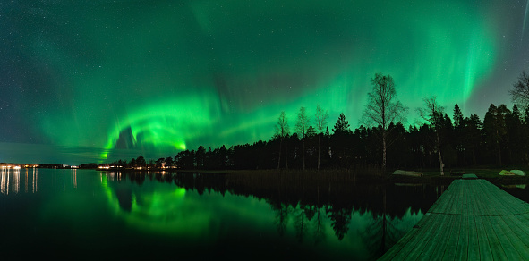 Spectacular night wide panorama strong green lights of dancing Aurora over Northern forest, reflection in lake, small boat, bridge. Stocksjo lake, Northern Sweden, Umea city