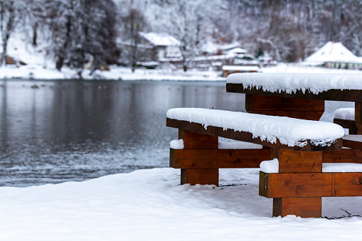 A wooden bench and a table near the lake surrounded by trees covered in the snow during the winter
