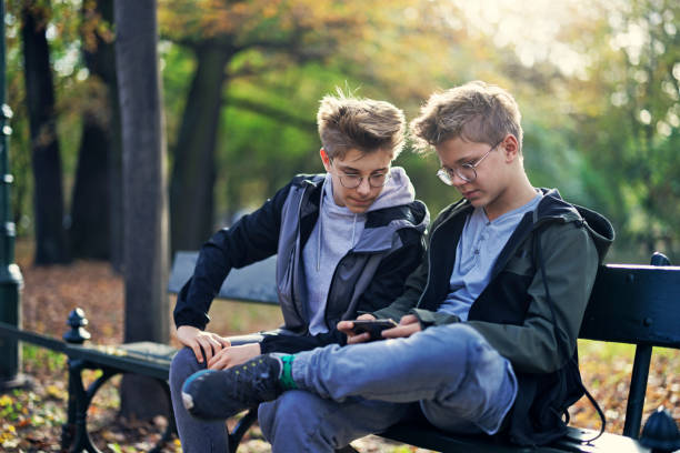Two teenage boys sitting on bench in public park and using smartphone on an autumn day.\nCanon R5