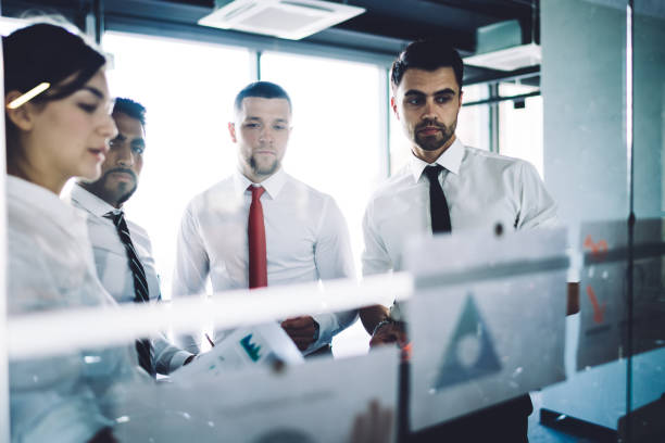 Group of male and female marketers collaborating during work day in office interior analyzing revenue statistics stick on glass board, diverse partners discussing graph charts during briefing