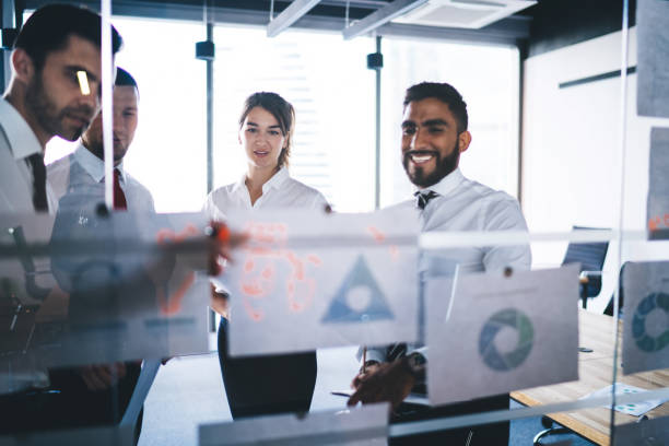 istock Multicultural group of corporate workers analyzing monetary gain of capital standing near glass stand with paper reports and searching negotiations for exchange experience, successful brainstorming 1438422175