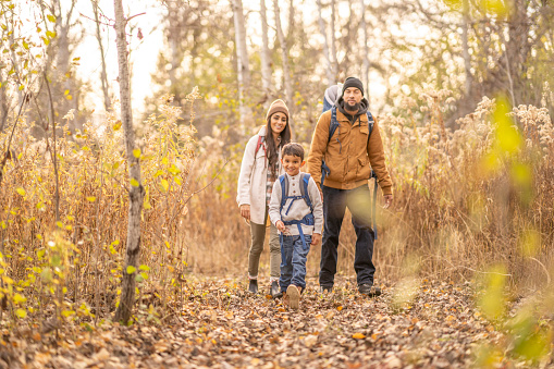 A Mother, Father and two children set out on a hike through the bare woods on a cool fall day.  The Father is carrying the baby in a backpack and the little boy has on a small backpack of supplies for their adventure. 
 They are each dressed warmly in layers as they crunch on the dry leaves underfoot.
