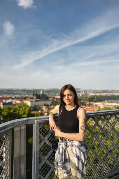 Portrait of a smiling young female in her 20´s posing with a slight smile at The Petřín Lookout Tower In Prague in a sunny day with some clouds and blurred background. She is wearing casual clothing and has one arm resting on the railing while looking at the camera.