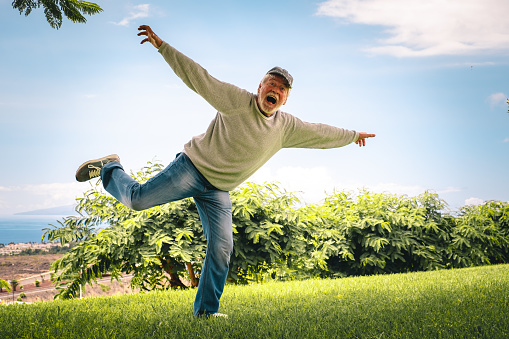 Joyful senior bearded man jumping in the meadow - caucasian grandfather in balance on one leg with open arms. Carefree and happy lifestyle for a retired people