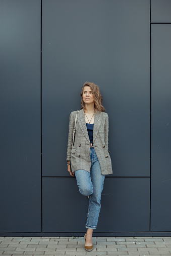 Vertical trendy confident, attractive, pensive blond businesswoman in gray jacket looking away and leaning on blue wall, posing in street. Fashion and modern outfit clothes and accessories. Copy space