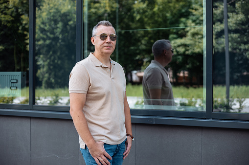 Confident, relaxed mature gray haired businessman in sunglasses, jeans and t-shirt stand near glass window of building with reflection. Urban street near park, stand with hands on belt, look at camera