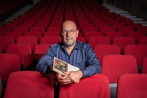 Confidence mature director sitting in an empty theater posing at camera with book