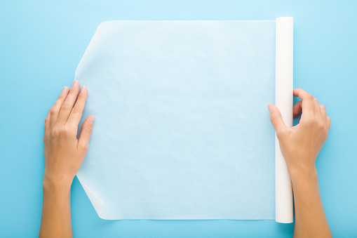 Young adult woman hands holding white roll of baking paper on light blue table background. Pastel color. Closeup. Point of view shot. Top down view.