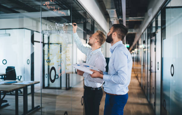 istock Smart caucasian 20s male employees concentrated on solving problem writing and drawing on glass in office, professional men colleagues cooperating and share strategy ideas together on job 1438443816