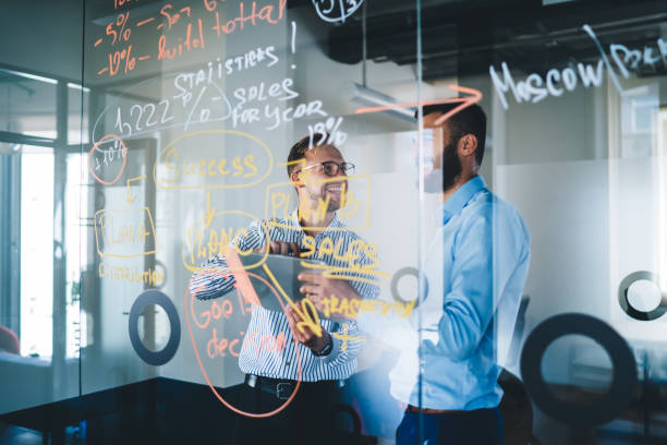 istock Cheerful partners smiling while collaborating near modern glass board with statistics planning for business marketing, entrepreneurs creating strategy indoors discussing web publication on gadget 1438443831