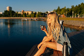 Relaxed blonde young woman holding cup with hot coffee from thermos sitting on beach by wooden pier on summer sunny morning.