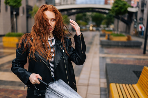 Portrait of frustrated pretty young woman with long red-hair standing with wet, disheveled hair after being caught in cold autumn rain, holding in hand broken umbrella, sad looking away.