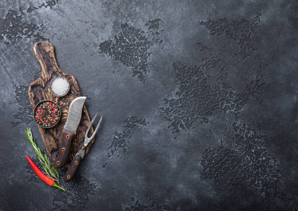 istock Fork and knife on chopping board with pepper and salt with rosemary on black background. 1438452231