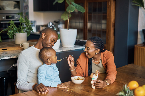 Eating, baby and parents with food in the kitchen of their house for breakfast, lunch or dinner. Young, happy and mother with smile while feeding a child at a table with love from dad in the morning