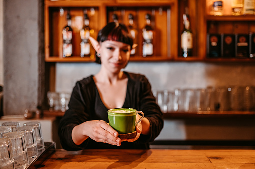 Young female barista wearing elf ears and serving a green cup of coffee for St, Patrick's day.