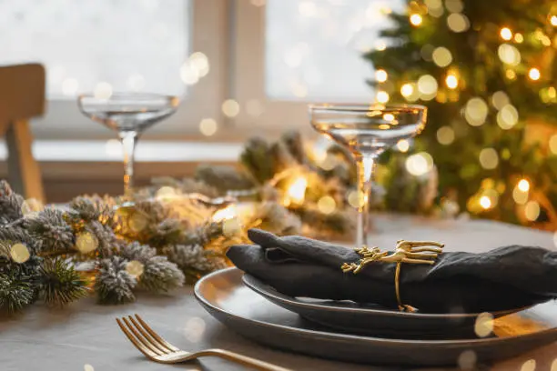 Christmas table setting with elegant empty gray plates decorated gold napkin ring near Xmas tree. Family holiday dinner at home. Close up.