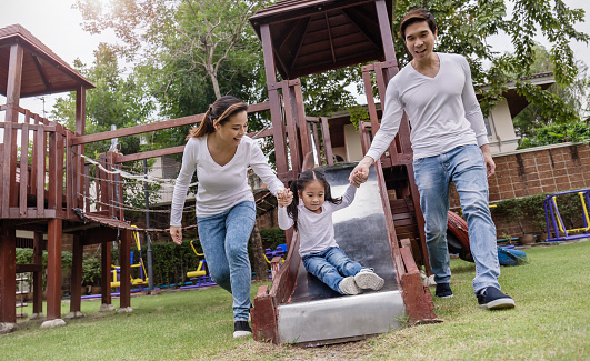Happy asian family mother father and daughter play slide. Asia parents and little toddler girl cuddling and hugging in the playground park. Happy family green spring or summer motherâs day together
