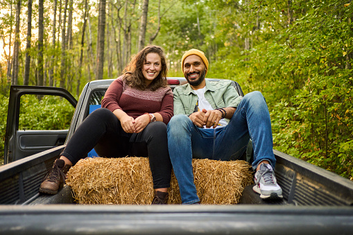 Portrait of a young couple smiling while sitting on hay bales in the back of a pick-up truck on a forest road