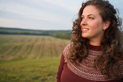 Smiling young woman with tousled hair looking at a scenic view at sunset while standing in a farm field