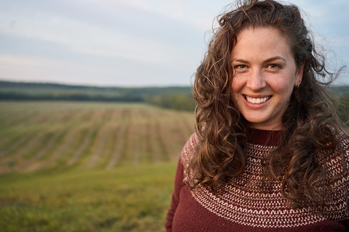 Portrait of a smiling young woman with tousled hair standing in a farm field at sunset