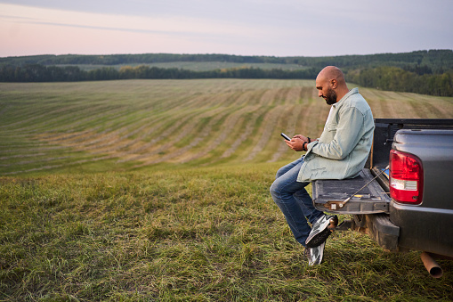 Young man texting on a phone while sitting in the back of his truck in a farm field at sunset