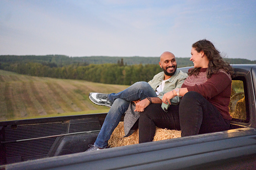 Young couple laughing while sitting on hay bales in the back of a pick-up truck in a farm field