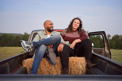 Portrait of a young couple smiling while sitting on hay bales in the back of a pick-up truck in a farm field