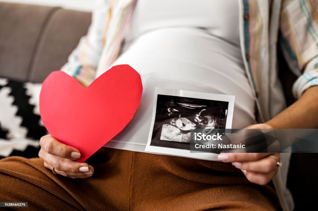 Unrecognizable pregnant woman holds heart shape and ultrasound image on pregnant belly A pregnant woman holds an ultrasound image 20-29 Years Stock Photo