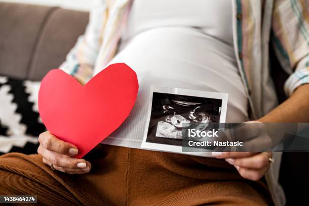 Unrecognizable Pregnant Woman Holds Heart Shape And Ultrasound Image On Pregnant Belly Stock Photo - Download Image Now