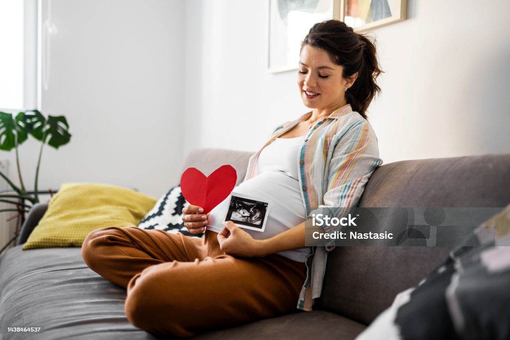 Smiling pregnant woman holds heart shape and ultrasound image on pregnant belly A pregnant woman holds an ultrasound image 20-29 Years Stock Photo
