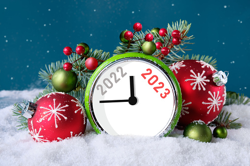 Year 2022 changes to 2023. Vintage alarm clock with fir twigs, cones, red trinkets and berries under snow. Dark green background with falling snow. Happy New Year 2023.