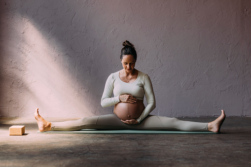 Portrait of a Caucasian woman sitting on a mat, stretching, holding her pregnant belly.
