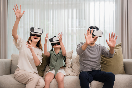 Moment happy of Asian family father, mother, and daughter wearing VR 3d or virtual reality glasses headset technology playing video game together on sofa living room at home. Relax innovation concept.