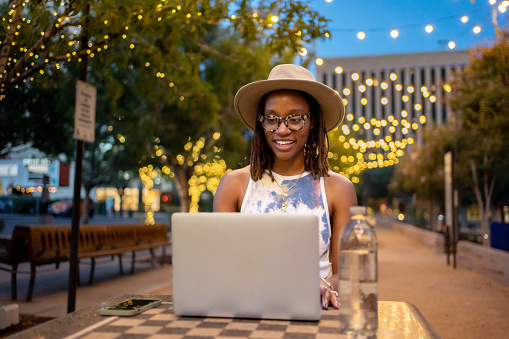 Pretty African-American Woman in Her Twenties Wearing a Hat in a Park in El Paso Texas Under Decorative Lighting Interacting on a Laptop on a Conference Call