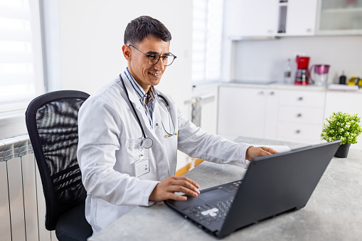 Beautiful cheerful young Caucasian male dentist sitting and writing reports on laptop