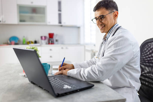 Cheerful doctor hand writing while looking at laptop stock photo