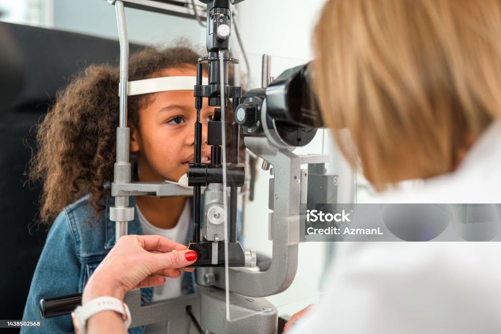 Curly Brown Hair Girl Sitting at Ophthalmologist Office Mature female optician doing optometry eye exam for African American child girl. Patient is wearing a jean jacket and two pony tails. 50-54 Years Stock Photo