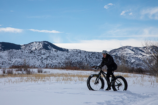 Morrison, Colorado, USA, February 15, 2020 Female fat tire mountain bike rider pedaling across snow covered field with Rocky Mountains in background and blue sky